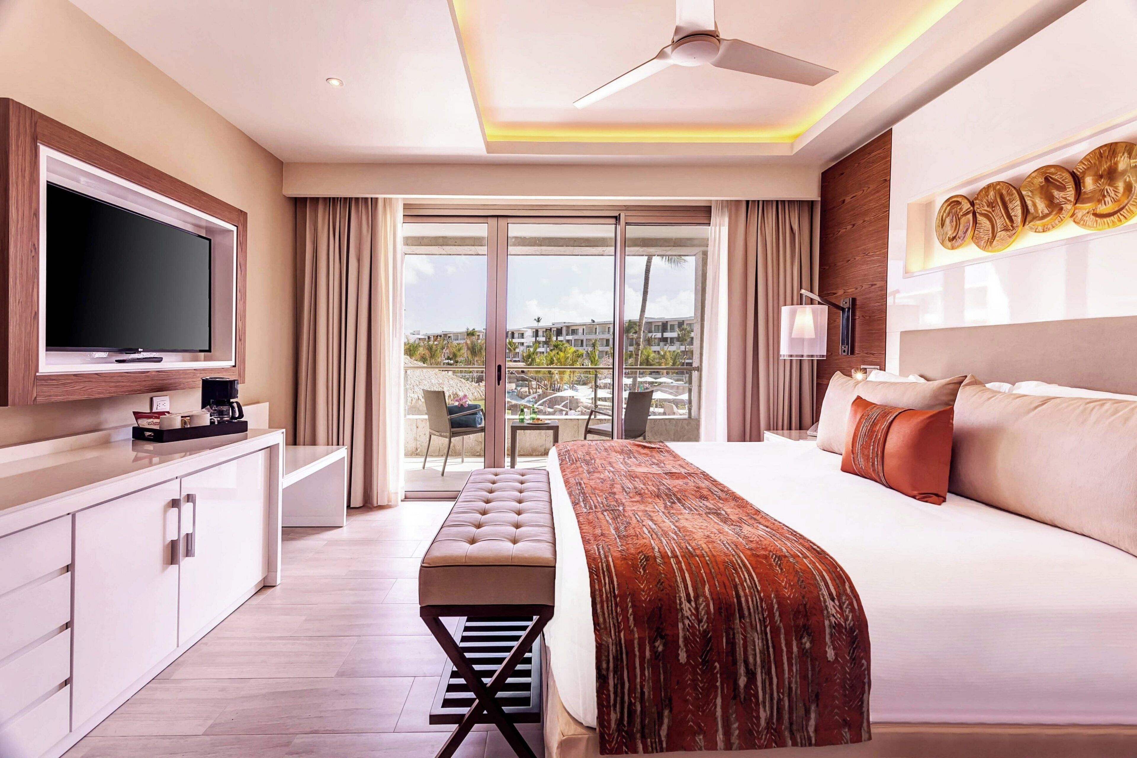 A modern hotel room with a large bed, a flat-screen TV, a desk, and a balcony with a view of buildings and palm trees.