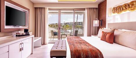 A modern hotel room with a large bed, a flat-screen TV, a desk, and a balcony with a view of buildings and palm trees.