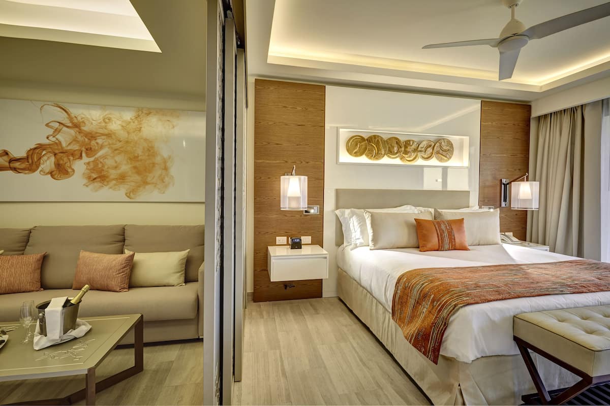 A modern hotel room with a large bed, a sofa, a small table with a bottle of champagne, and a wall art piece.