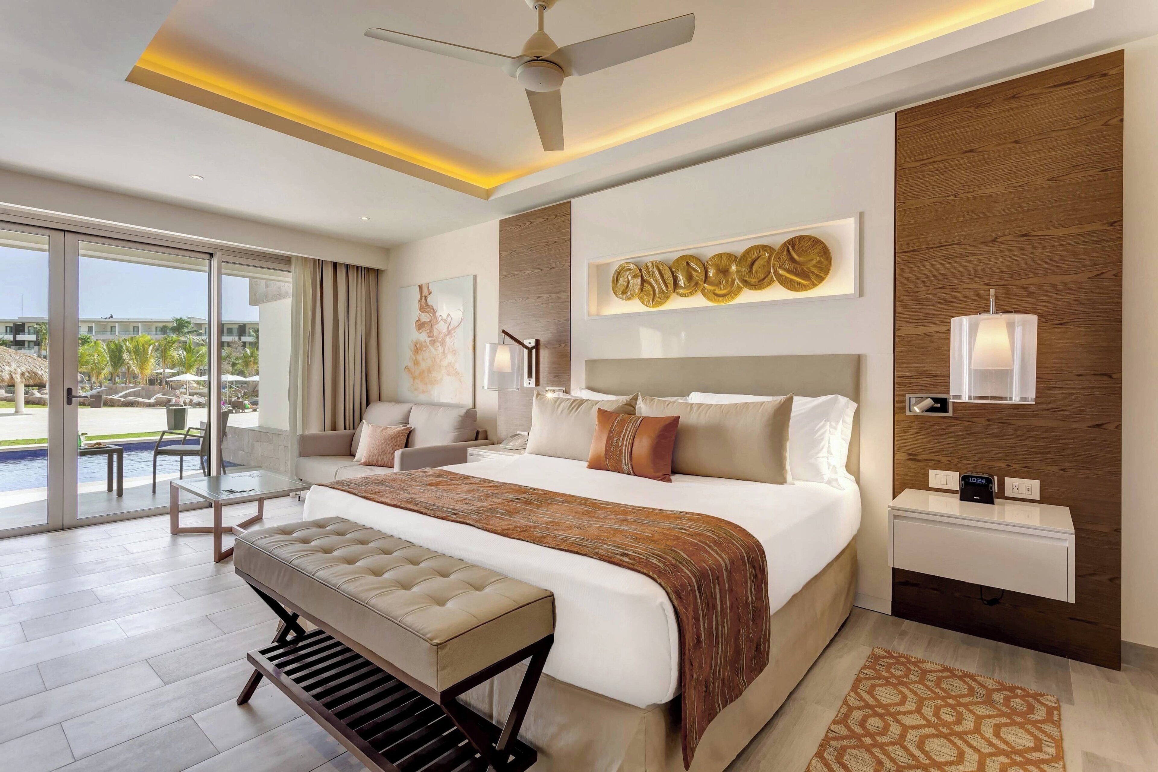 A modern hotel room with a large bed, a bench, a desk, and a view of the pool area.