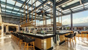 Un'ampia zona bar con soffitti alti e un design moderno, caratterizzata da un lungo bancone con sgabelli e una vasta selezione di bottiglie.