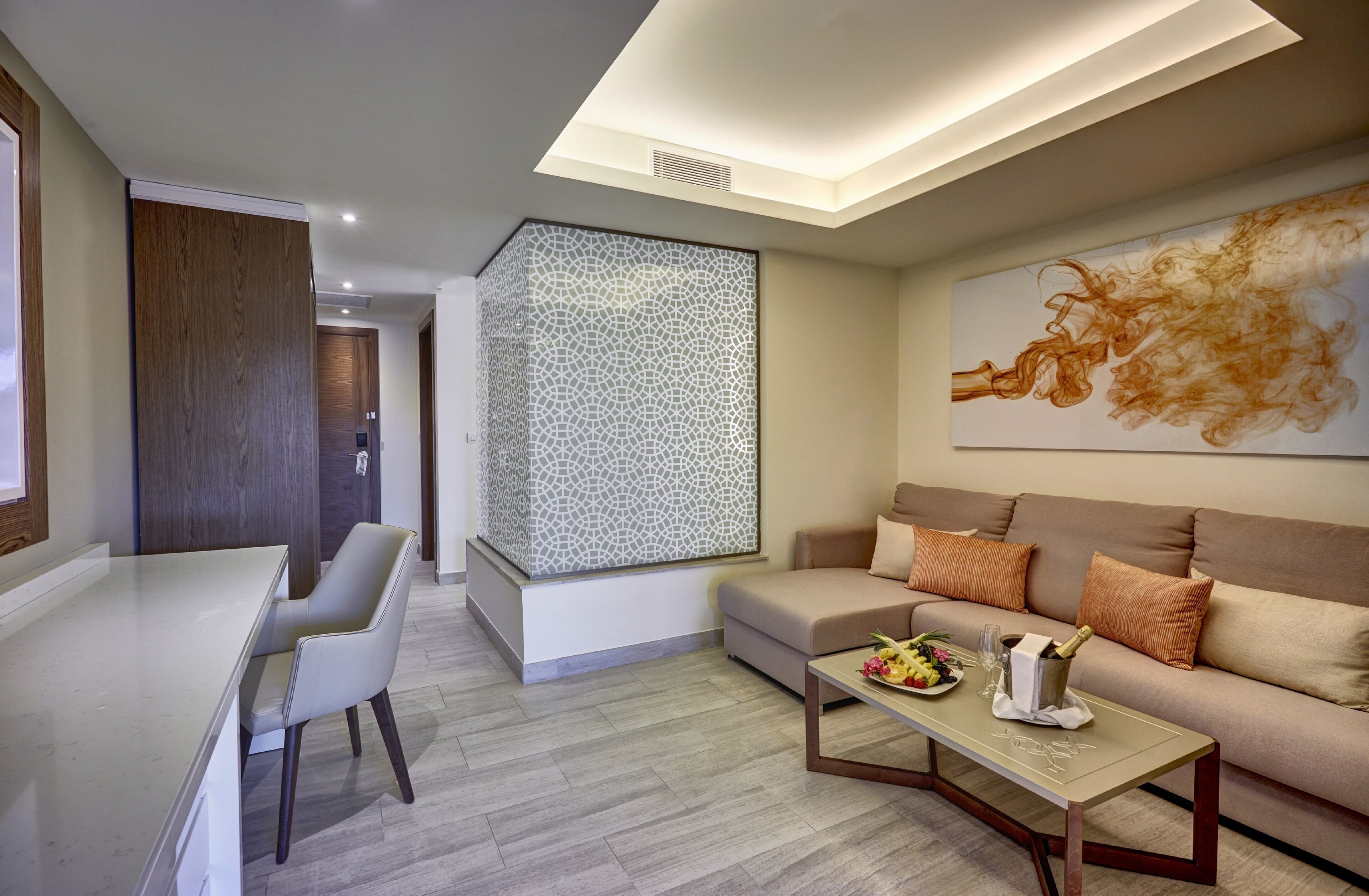 A modern living room with a sofa, a coffee table with a bottle and flowers, a dining area with a chair, and a decorative wall panel.
