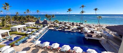 A resort pool area with multiple lounge chairs and umbrellas, a central swimming pool, and a view of the ocean.