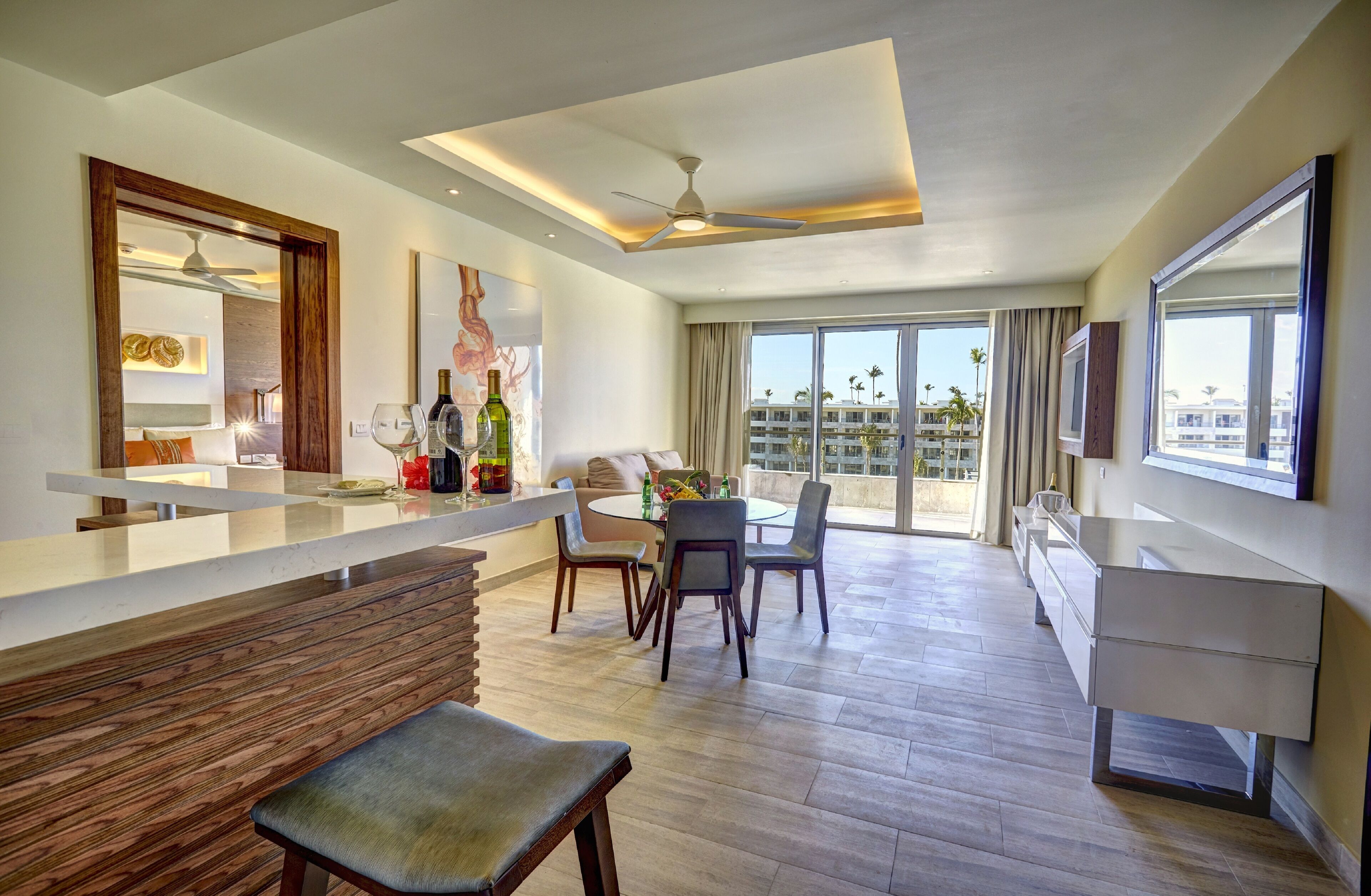 A modern hotel room with a dining area, a balcony with a view of palm trees, and a large window.