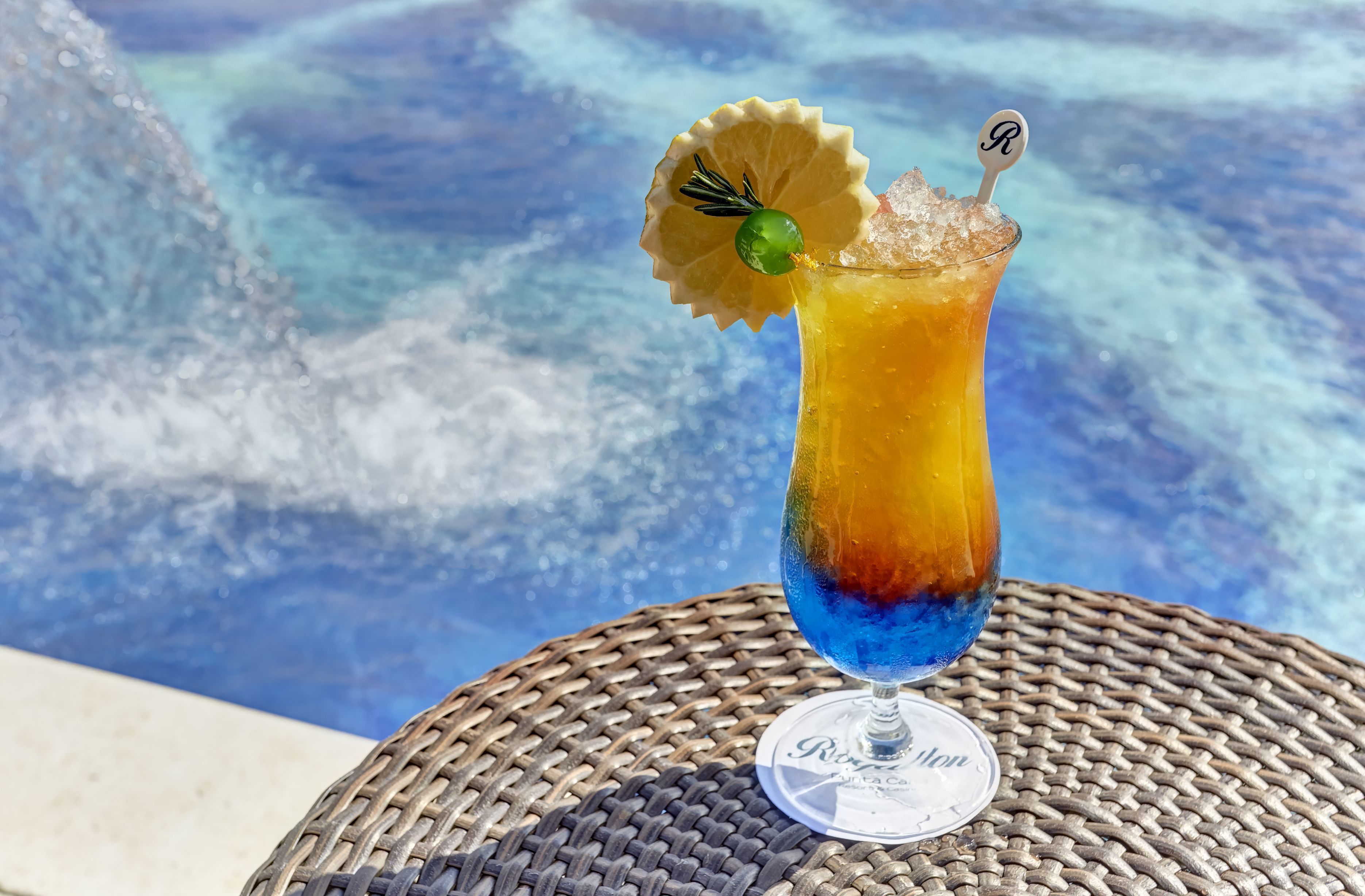 A cocktail with a layered effect, garnished with a lemon slice and a green sphere, on a glass with a coaster labeled "Riviera."