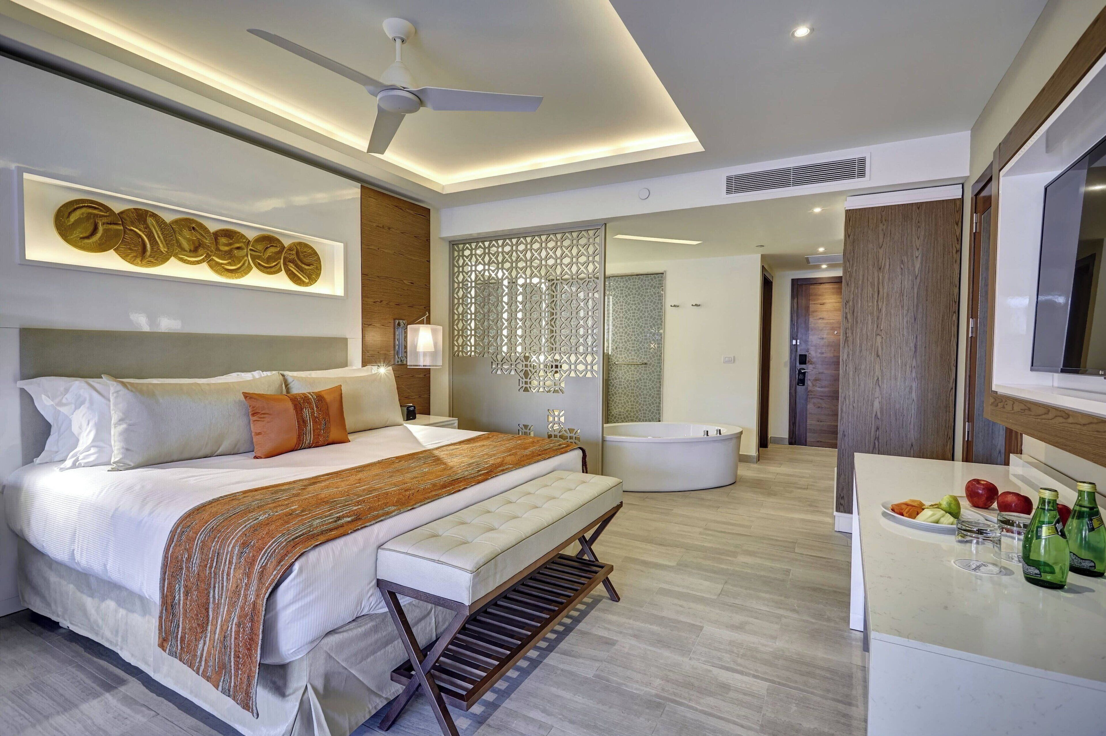 Une chambre d’hôtel moderne dotée d’un grand lit, d’une table de chevet, d’une salle de bain avec baignoire et d’un espace pour le petit-déjeuner où sont servis des fruits et des boissons.