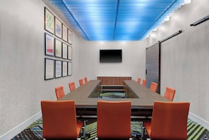 Sala de conferência com uma mesa comprida, cadeiras laranja, uma TV de tela plana e quadros nas paredes.