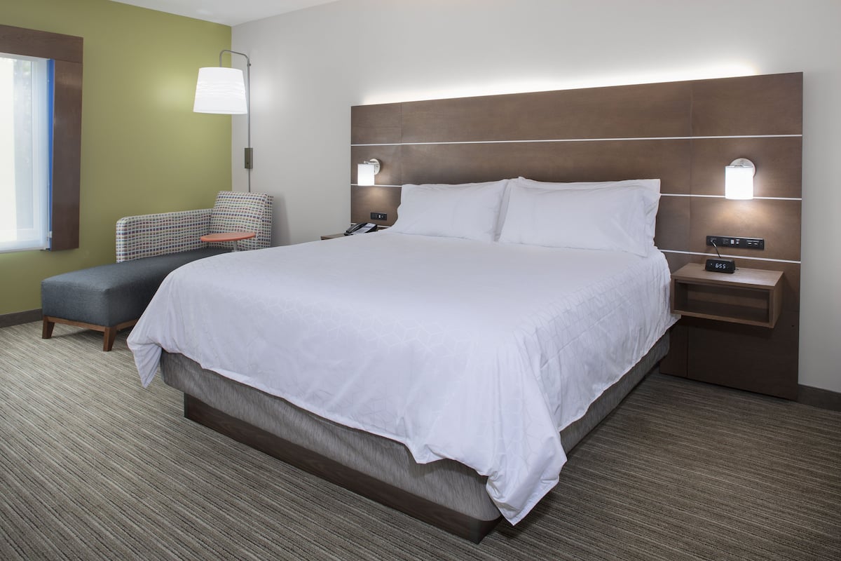 A hotel room with a large bed, a bedside table with a lamp, a chair, and a small table.