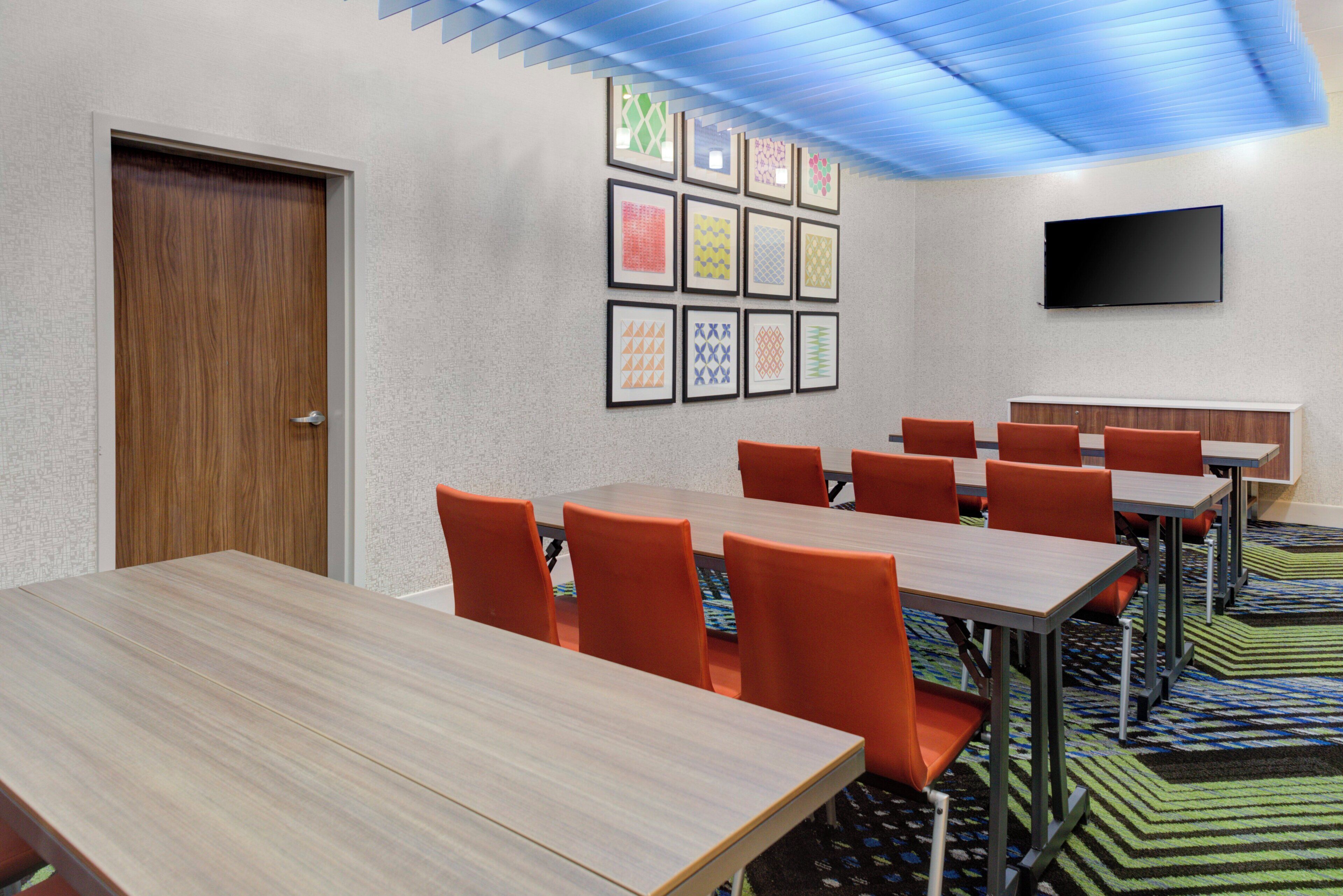 Un salón de conferencias con una mesa de madera, sillas naranja, un televisor de pantalla plana y cuadros en la pared.
