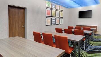 Un salón de conferencias con una mesa de madera, sillas naranja, un televisor de pantalla plana y cuadros en la pared.