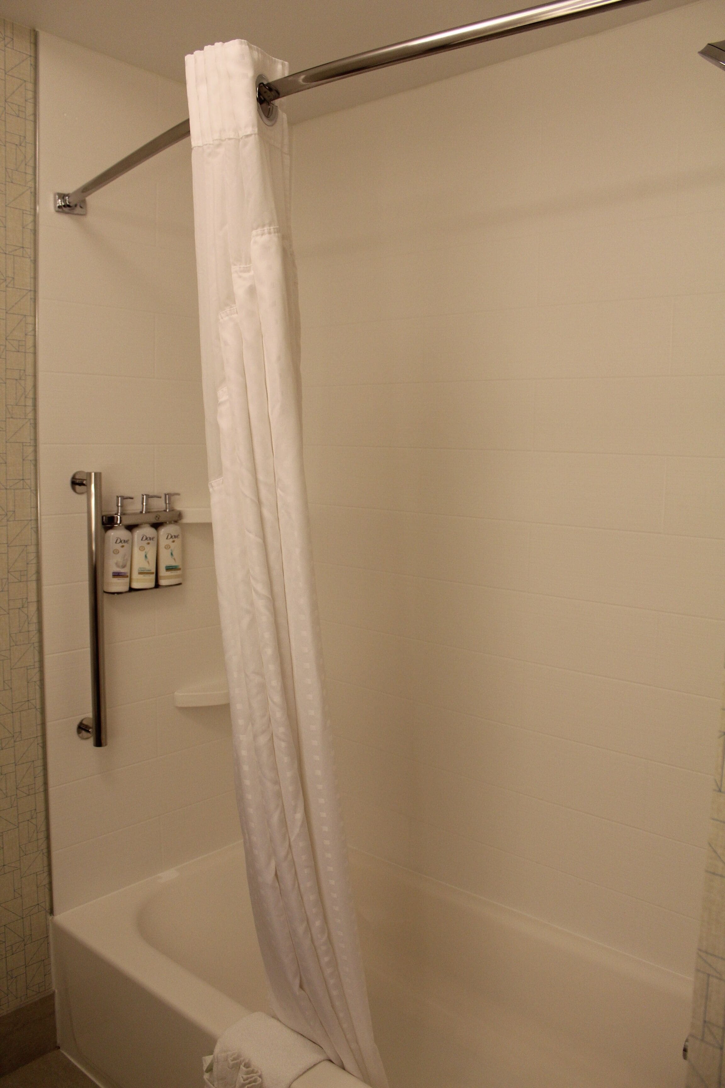 A bathroom with a white bathtub, a shower curtain, and a handrail. There are three bottles of Dove body wash on a shelf.