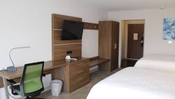 A hotel room with a wooden desk, a green office chair, a bed with white bedding, a television mounted on the wall, and a door leading to another room.