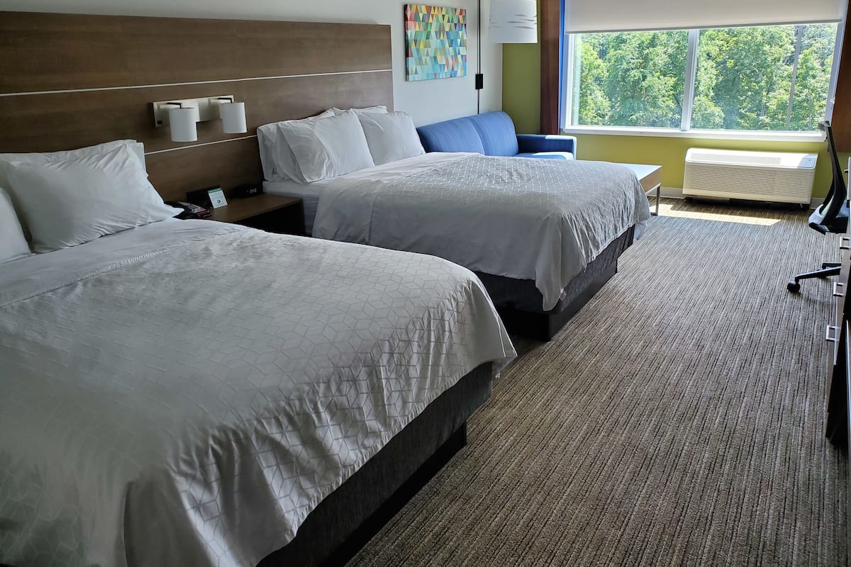 A hotel room with two beds, a desk, a chair, a lamp, and a window with a view of trees.