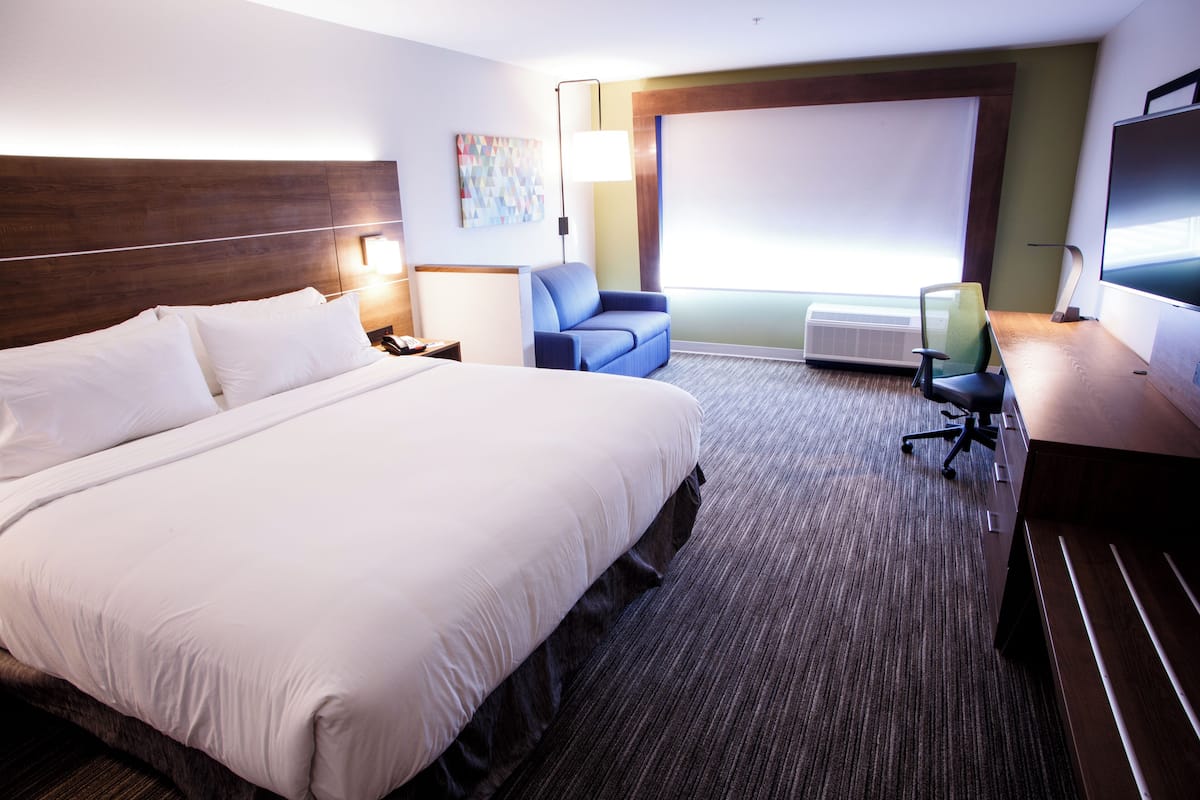 A hotel room with a large bed, a desk, a chair, a flat-screen TV, and a window with blinds.