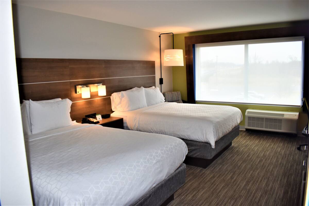 A hotel room with two beds, a nightstand, a lamp, and a large window.