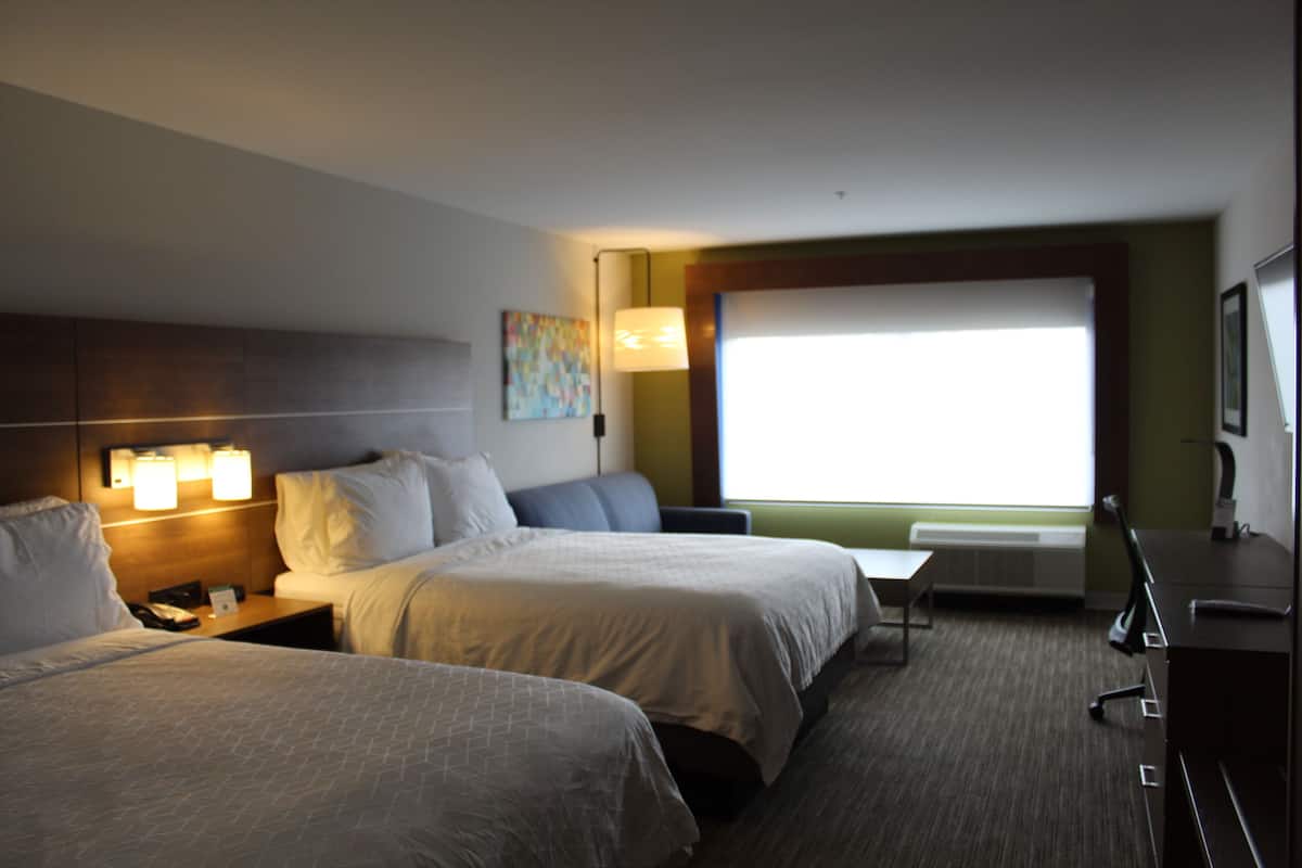 A hotel room with two beds, a desk, a chair, a lamp, and a window with blinds.