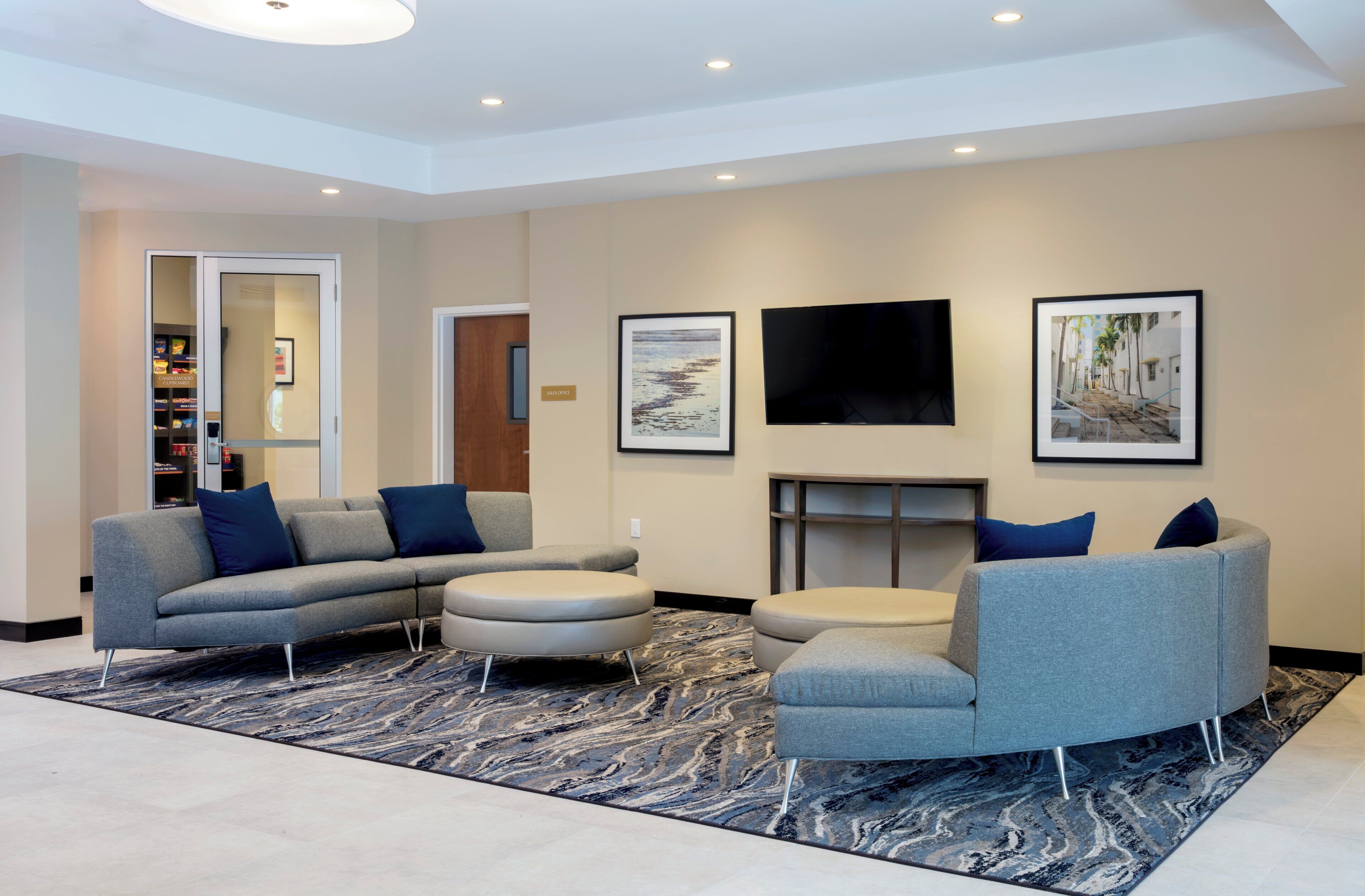 A modern living room with a sectional sofa, a round ottoman, a grey armchair, a flat-screen TV, and framed artwork on the walls.