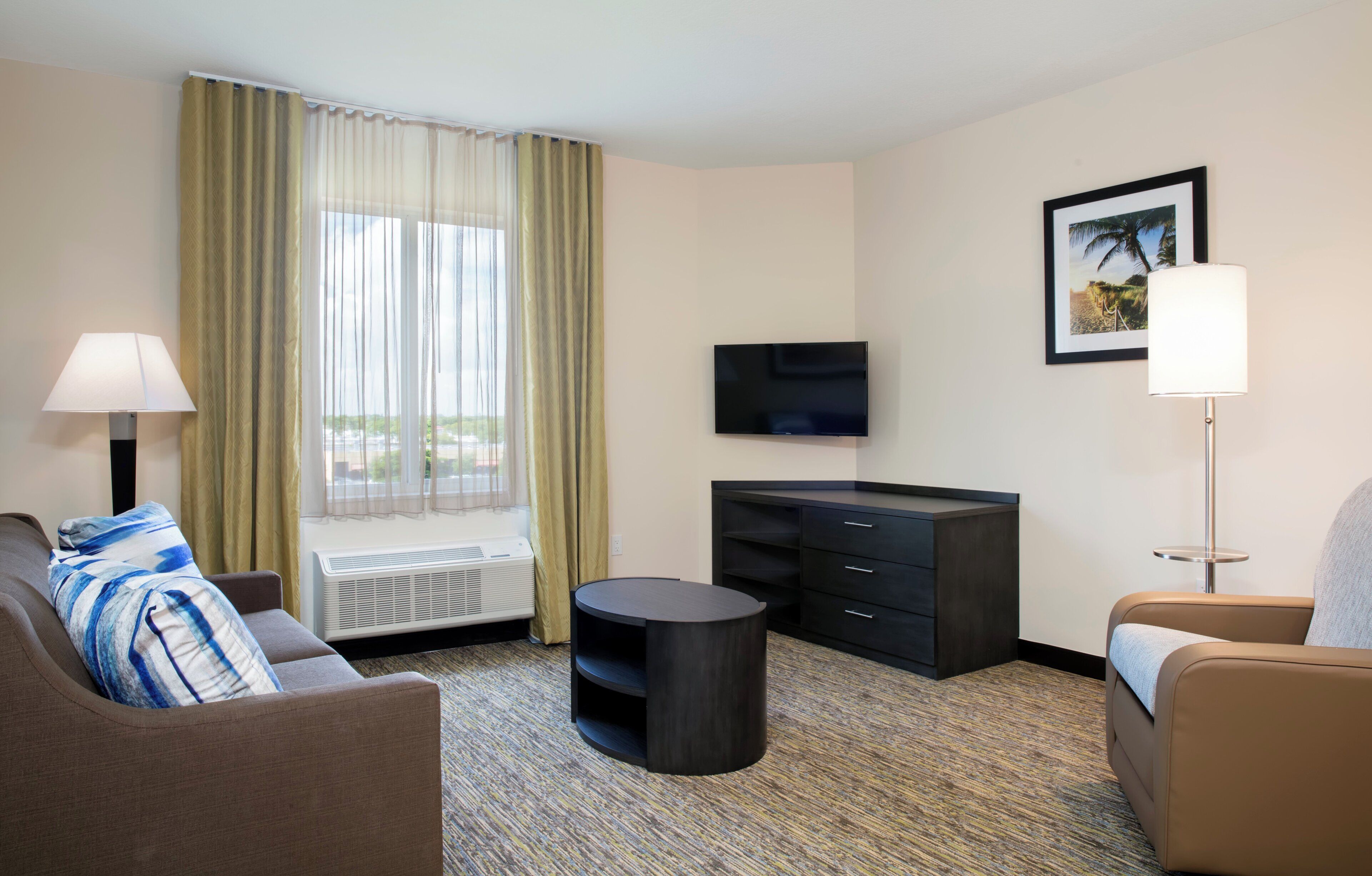 Une chambre d’hôtel avec un canapé, une petite table ronde, un téléviseur, une armoire et une fenêtre avec des rideaux.