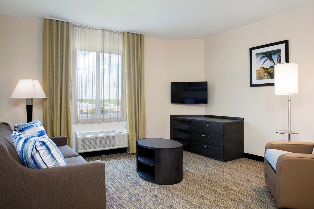Une chambre d’hôtel avec un canapé, une petite table ronde, un téléviseur, une armoire et une fenêtre avec des rideaux.