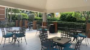 An outdoor patio area with tables and chairs, a grill, and a view of greenery.