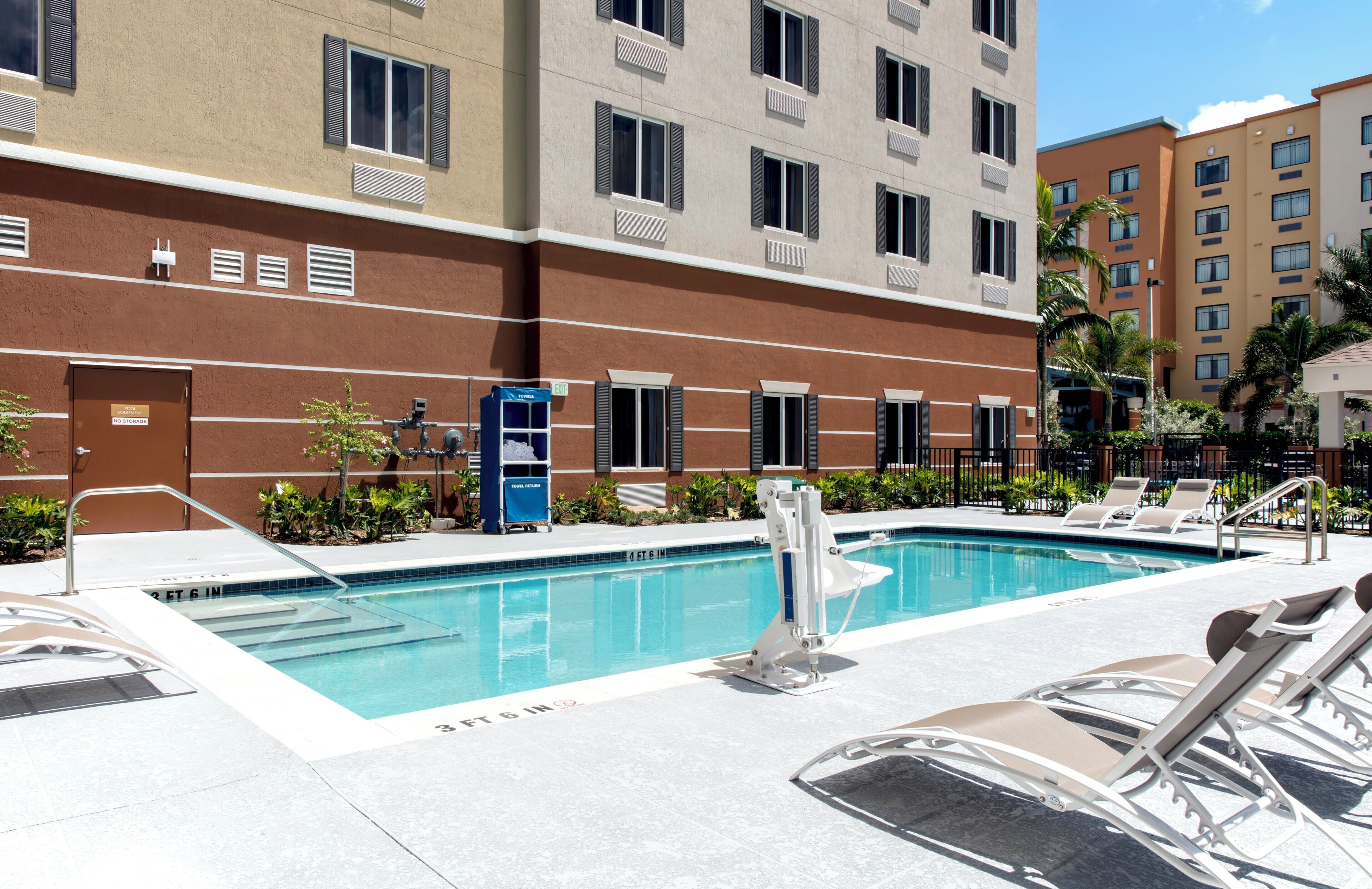 Área da piscina com piscina límpida, espreguiçadeiras e um edifício com várias janelas e uma porta com a inscrição 'Devolução de Toalhas'.