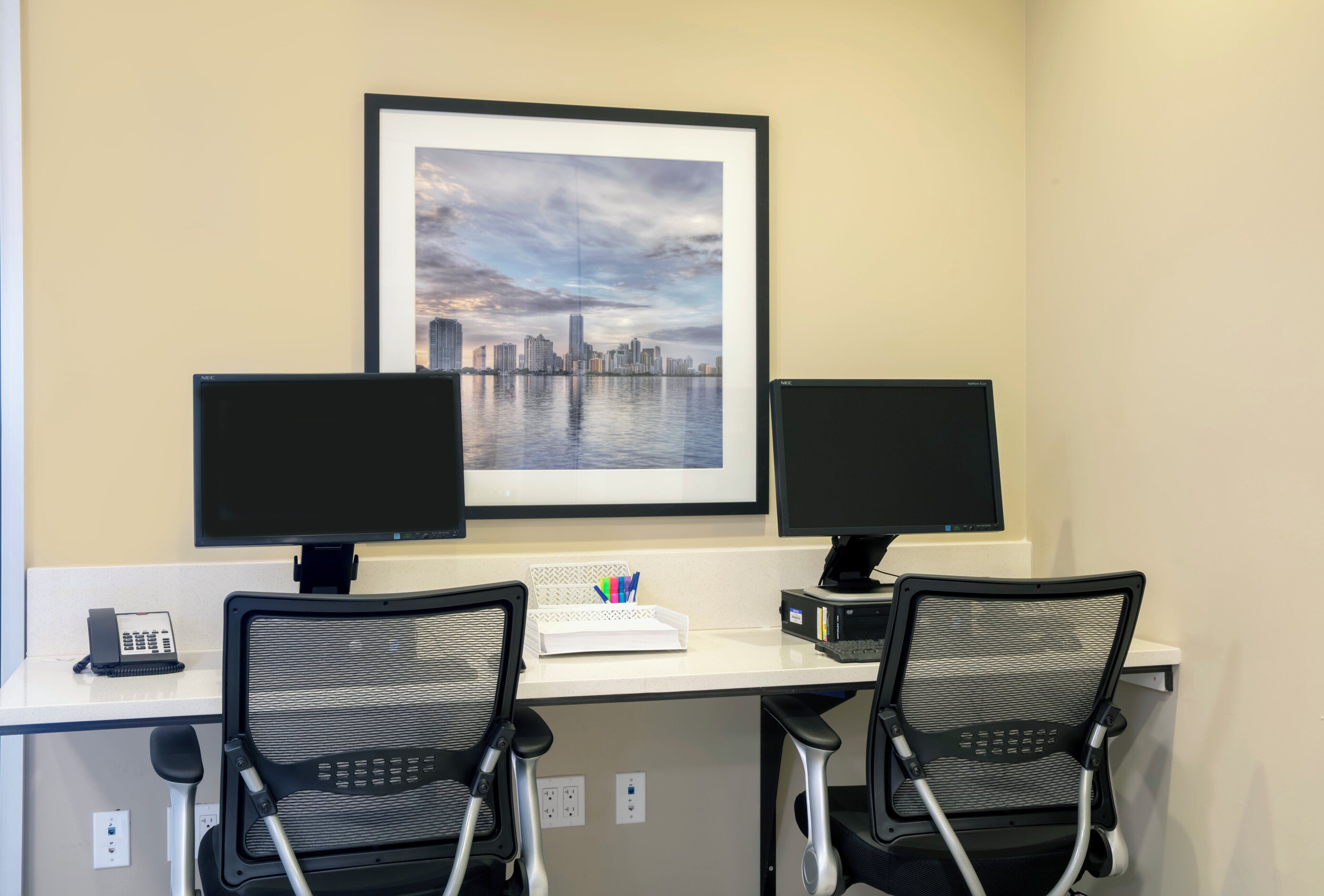 A workspace with two monitors, a framed cityscape, a phone, and two office chairs.