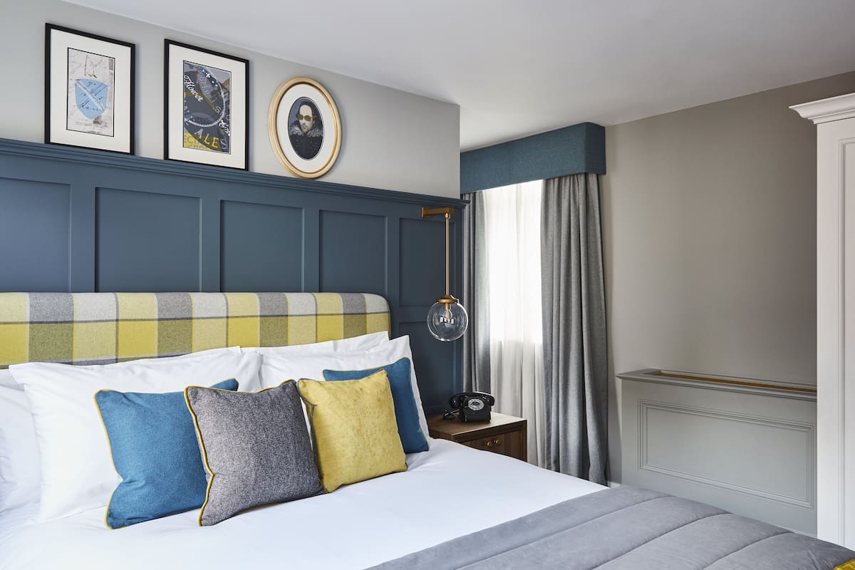 A neatly arranged bedroom with a bed, pillows, a bedside table with a telephone, and framed pictures on the wall.