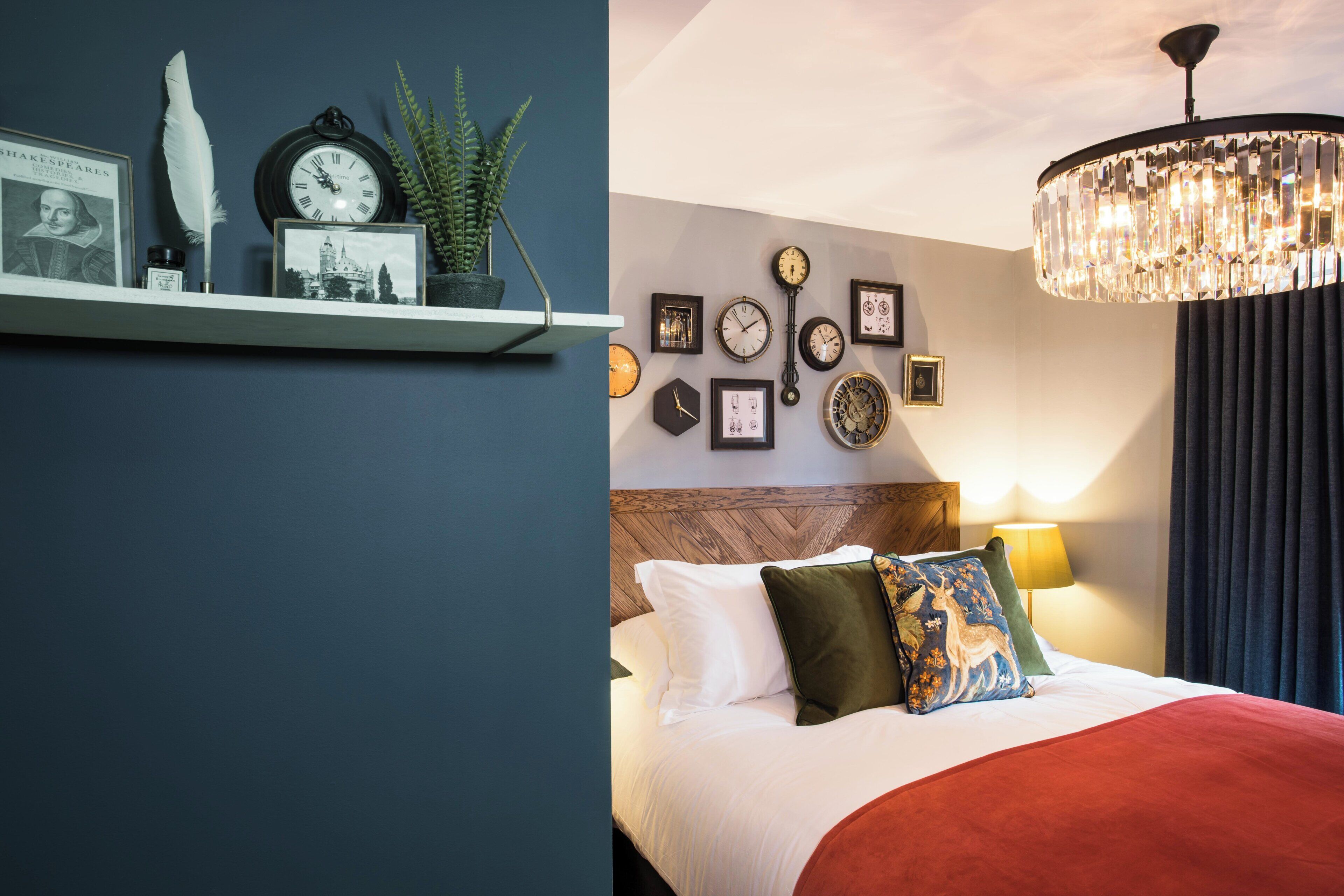 A bedroom with a bed, multiple clocks on the wall, a chandelier, and a shelf with decorative items.