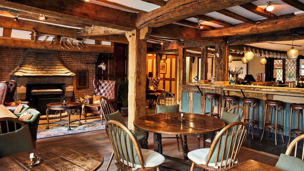 Un intérieur rustique avec une cheminée en briques, des poutres en bois, un coin bar et différents espaces pour s’asseoir.