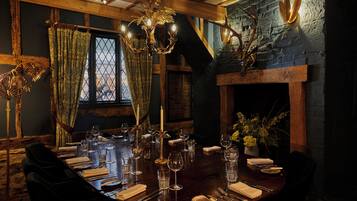 A dining room with a long table set for a meal, a chandelier, and a fireplace with antlers.