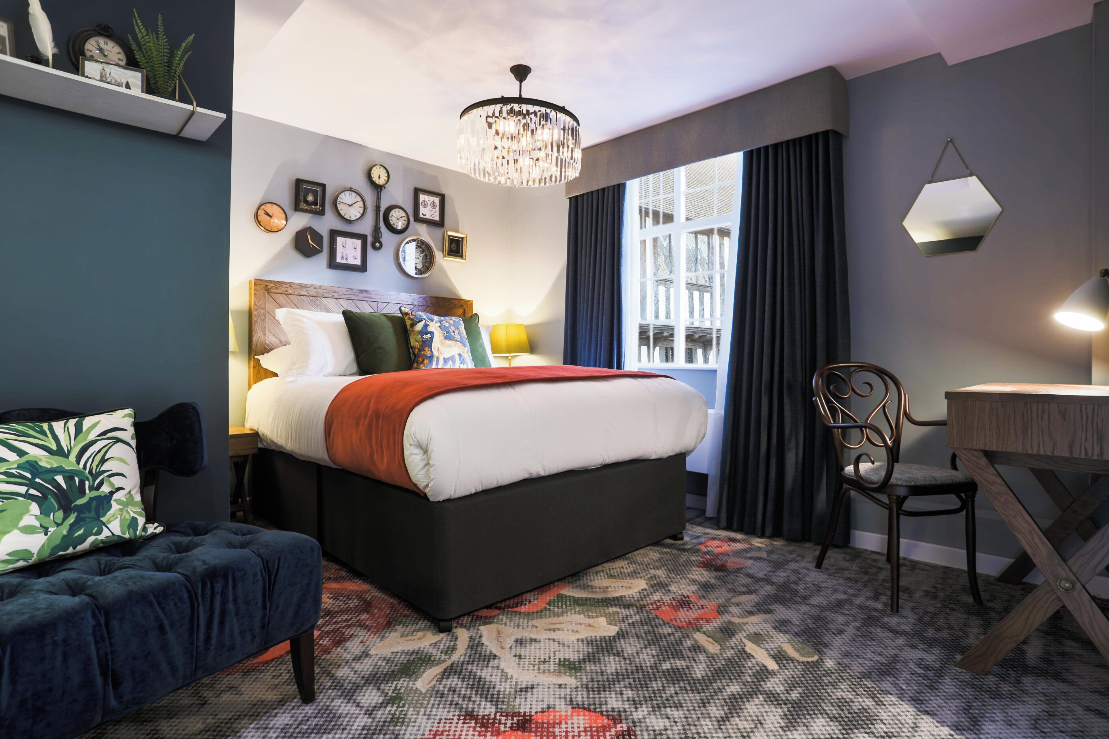 A hotel room with a bed, a desk, a chair, a window with curtains, a chandelier, and wall clocks.