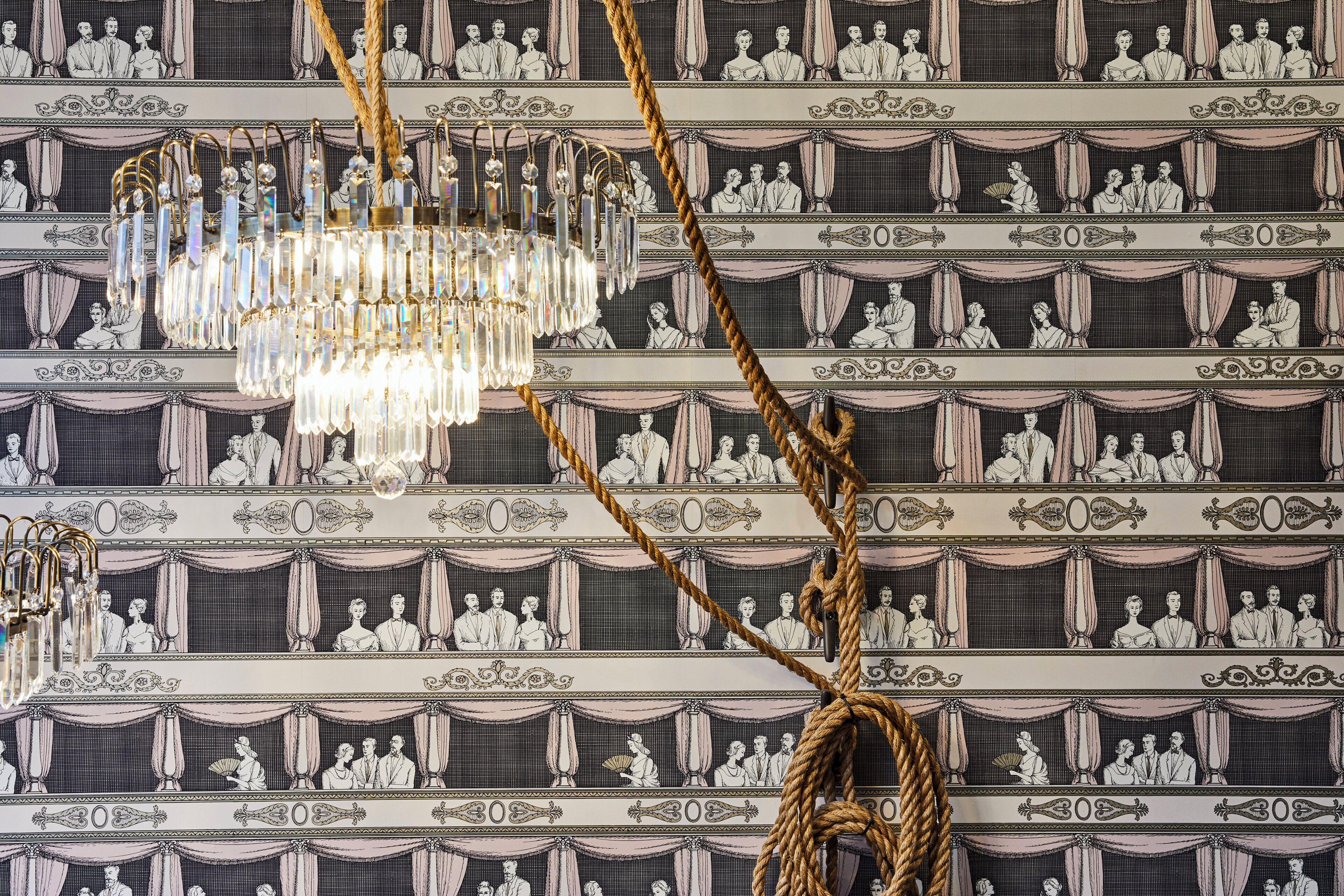 A chandelier with multiple crystal elements and a golden rope decoration against a wallpaper with a repeated pattern of figures and the number '300'.