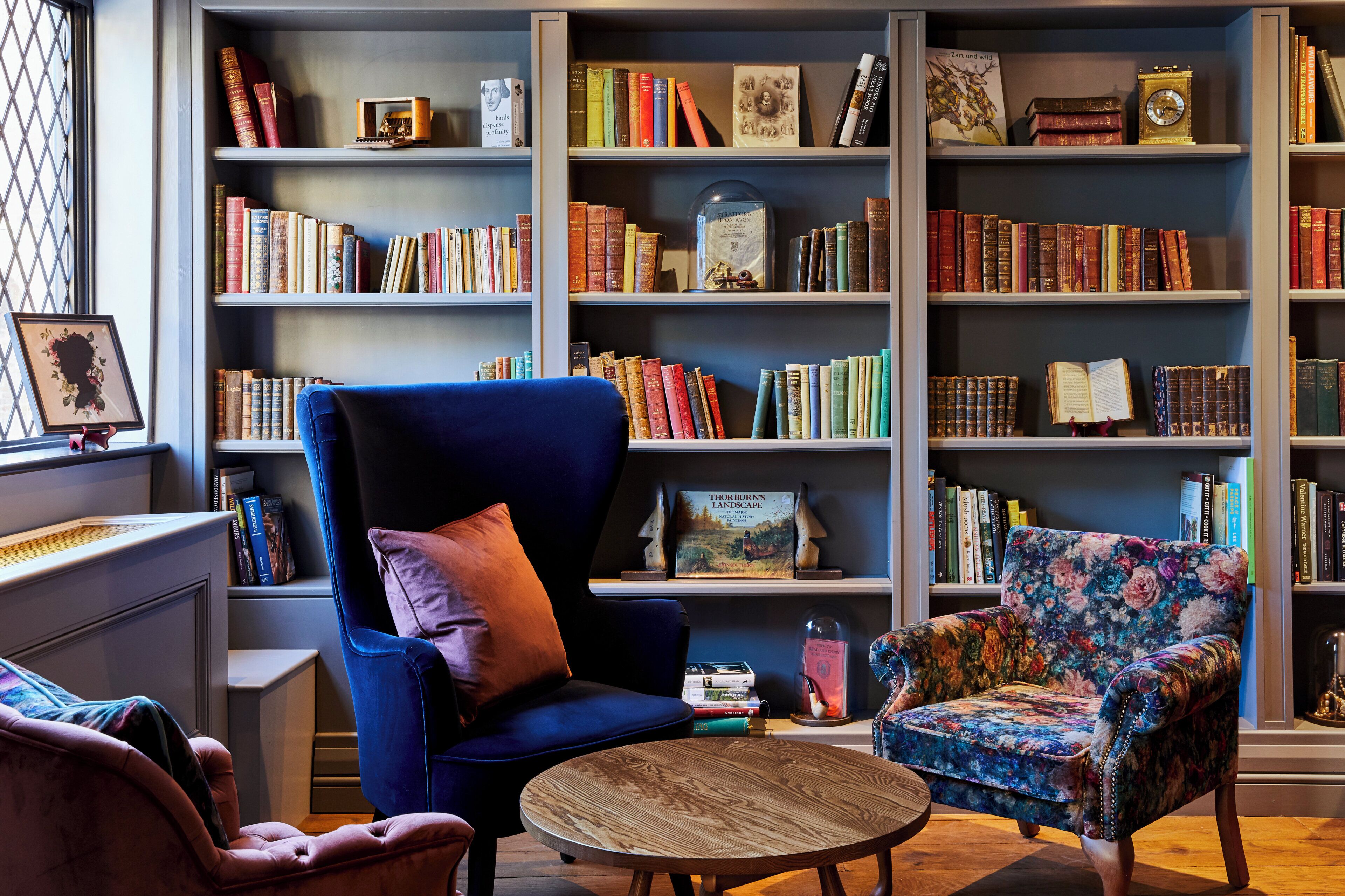 A library with bookshelves, a blue armchair, a round wooden table, and a floral armchair.