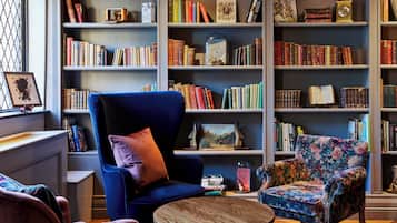 A library with bookshelves, a blue armchair, a round wooden table, and a floral armchair.