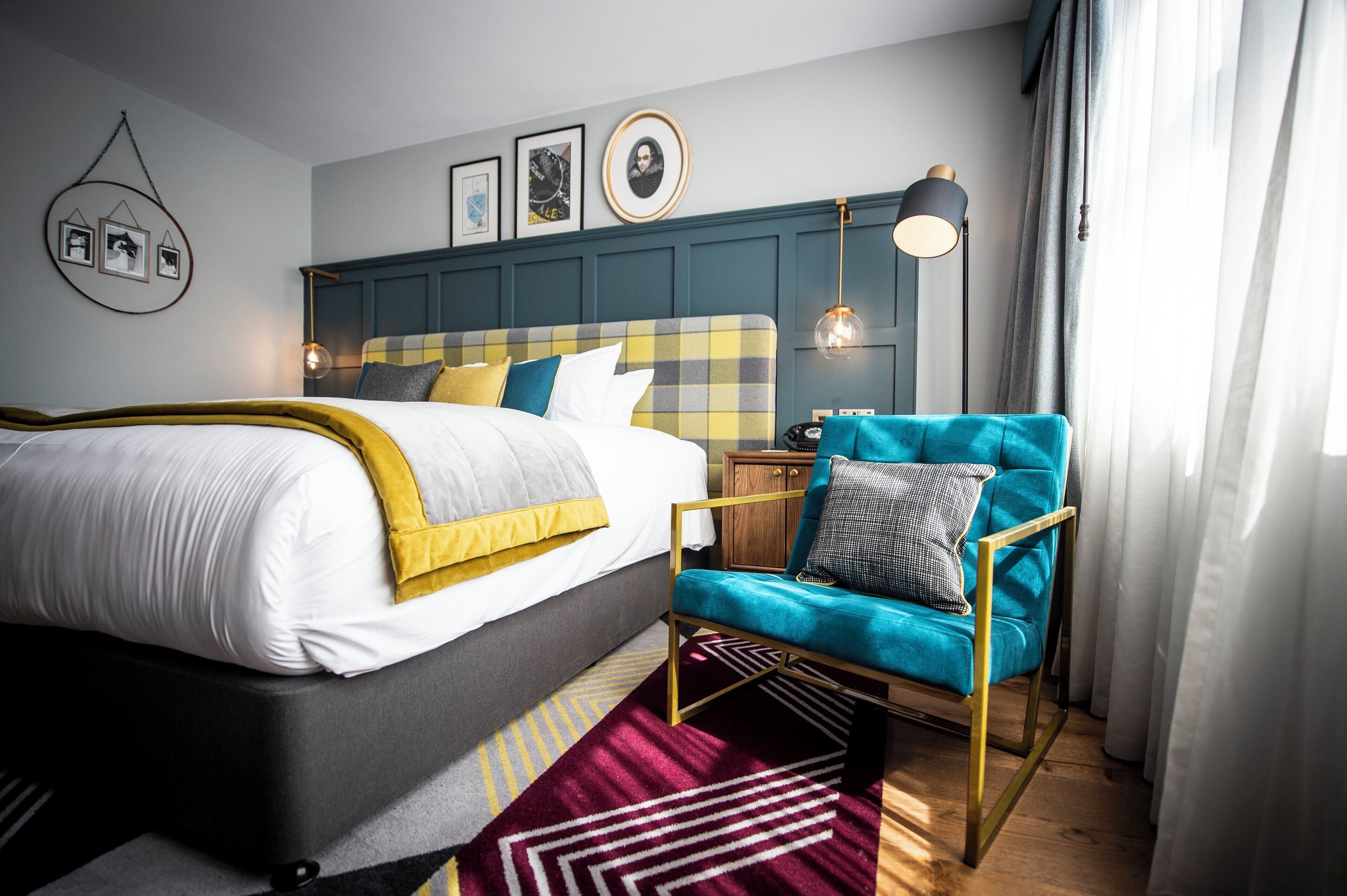 A neatly arranged bedroom with a bed, a blue armchair, a bedside table, and a wall-mounted lamp.