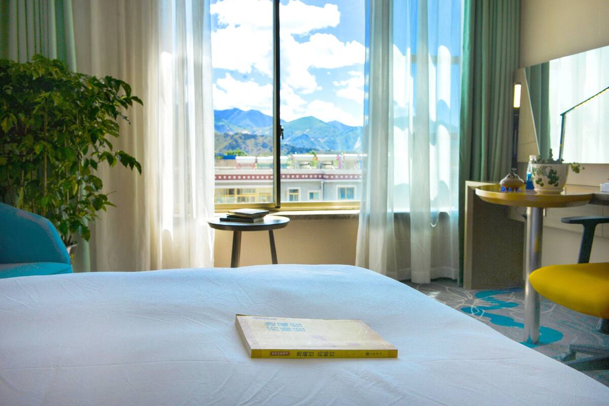 A hotel room with a bed, a small table with a book, a chair, a potted plant, and a view of mountains through the window.