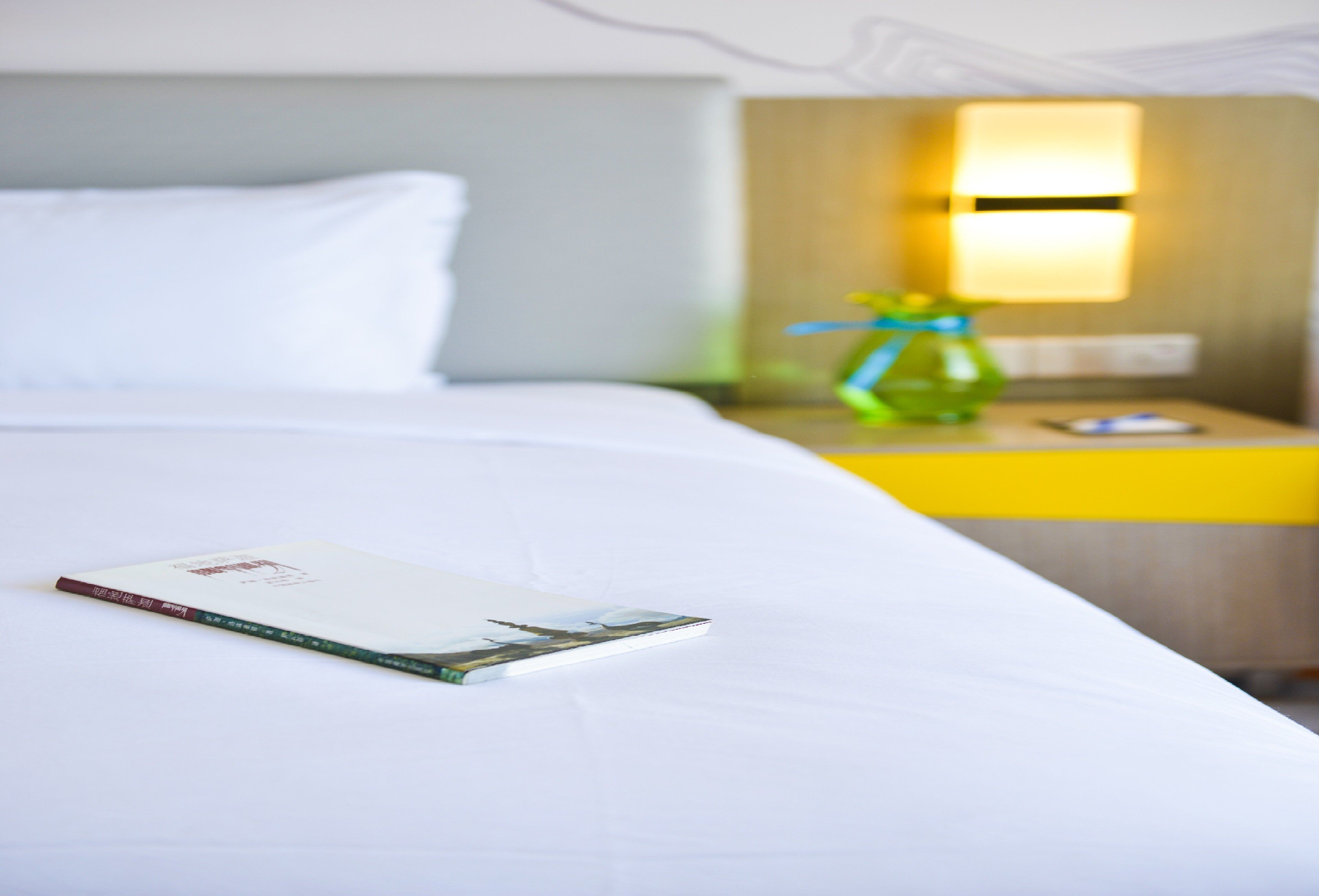 A neatly made bed with a pillow and a book on it, a bedside table with a lamp and a bottle, and a yellow nightstand.