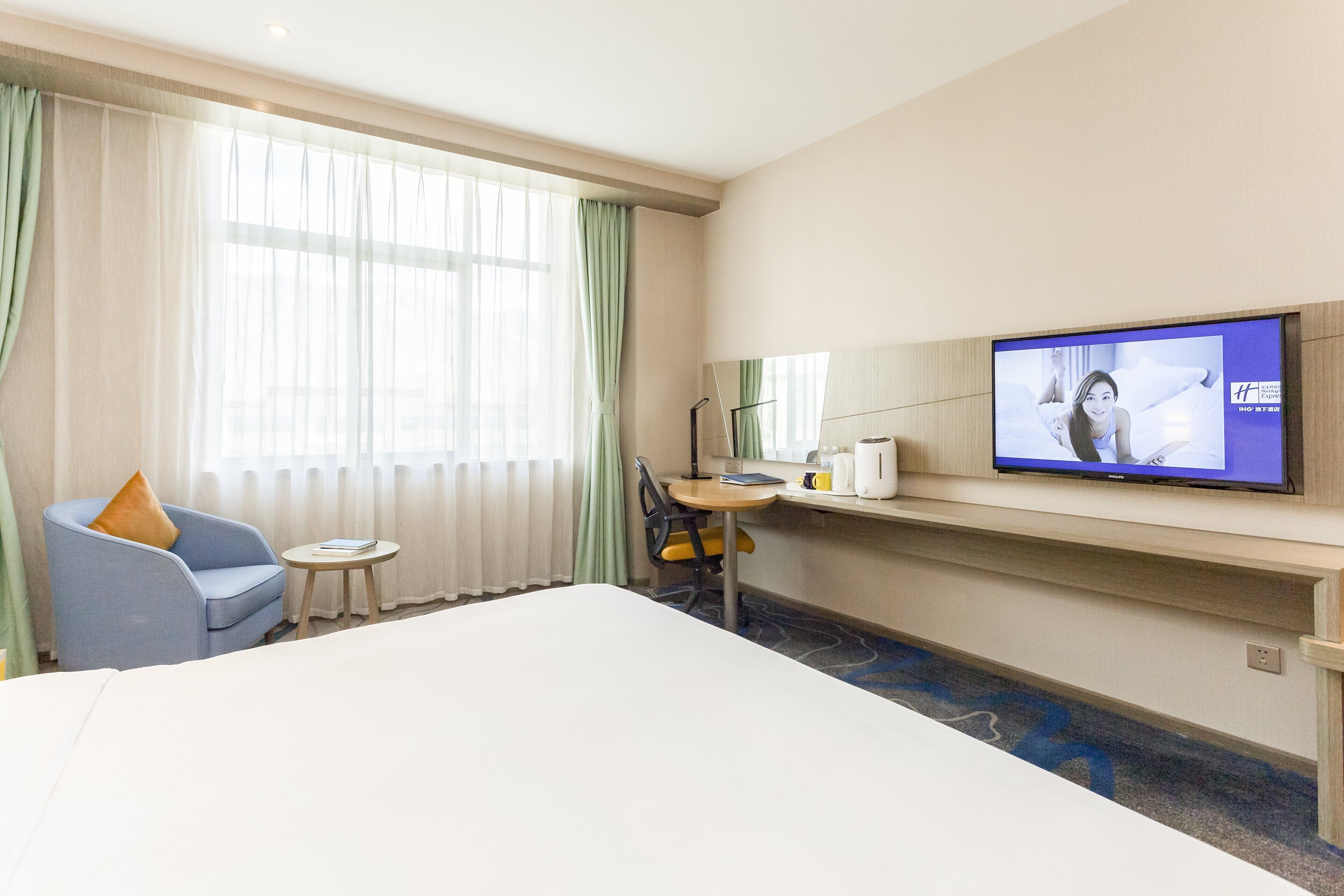 Une chambre d’hôtel avec un grand lit, un téléviseur fixé au mur, une petite table avec une chaise et une fenêtre avec des rideaux légers.