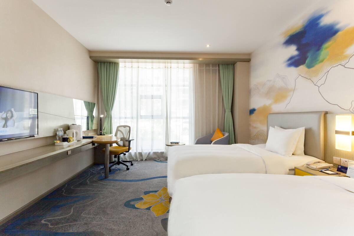 A hotel room with two beds, a desk with a chair, a TV, and a large window with curtains.