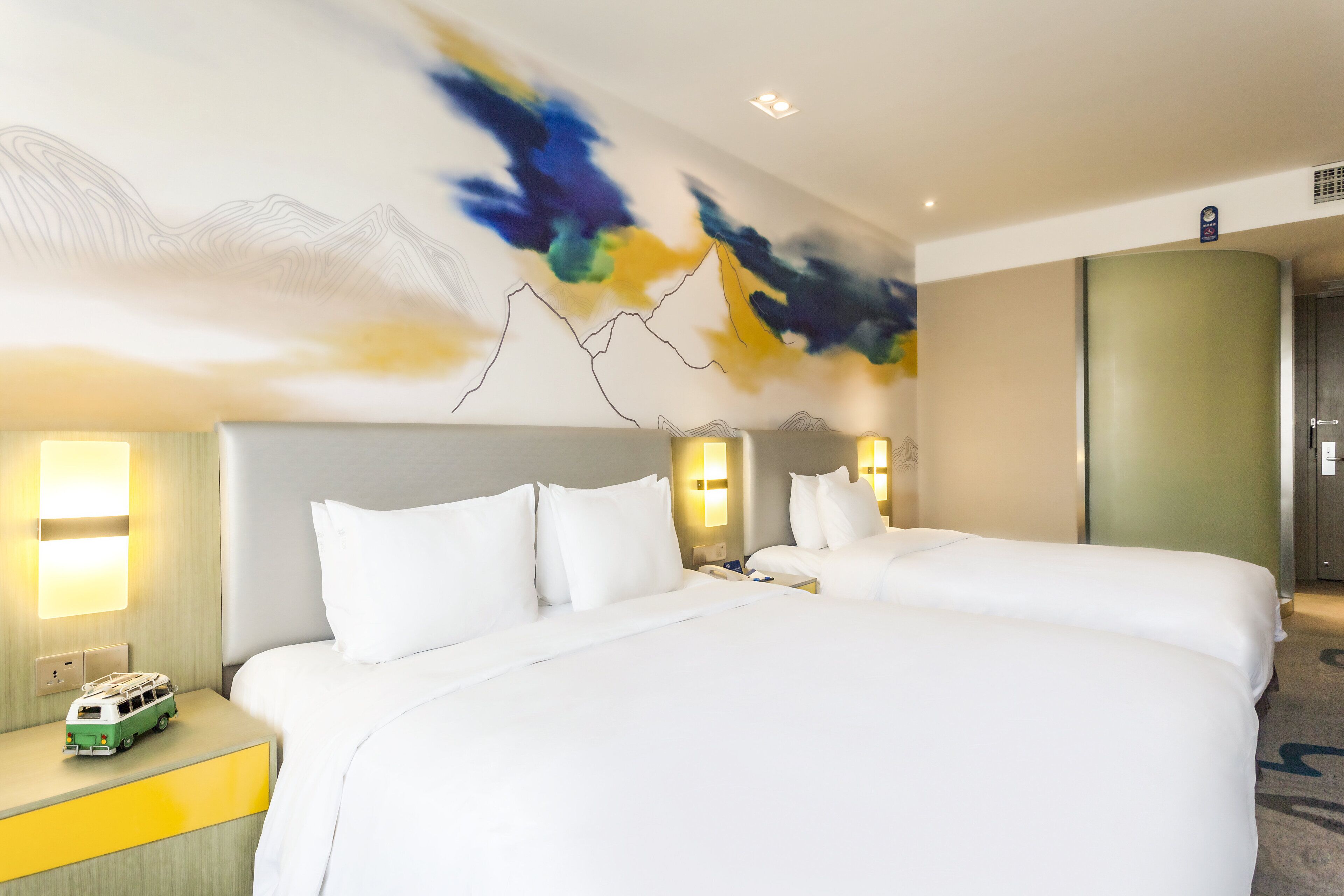 Habitación de hotel con dos camas, una mesita con un coche de juguete y una pared con arte abstracto.