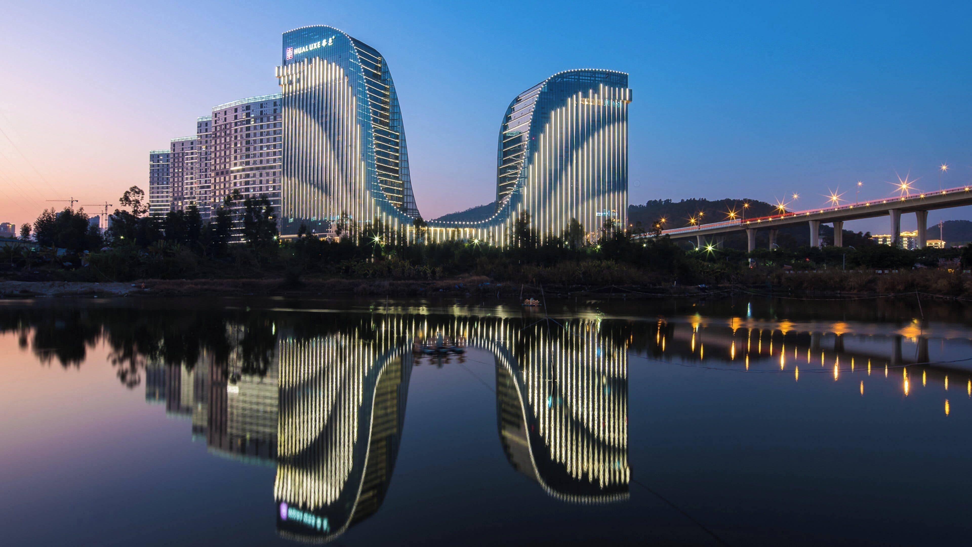 Un bâtiment moderne à la façade incurvée, qui se reflète dans l’eau, avec un pont en arrière-plan.