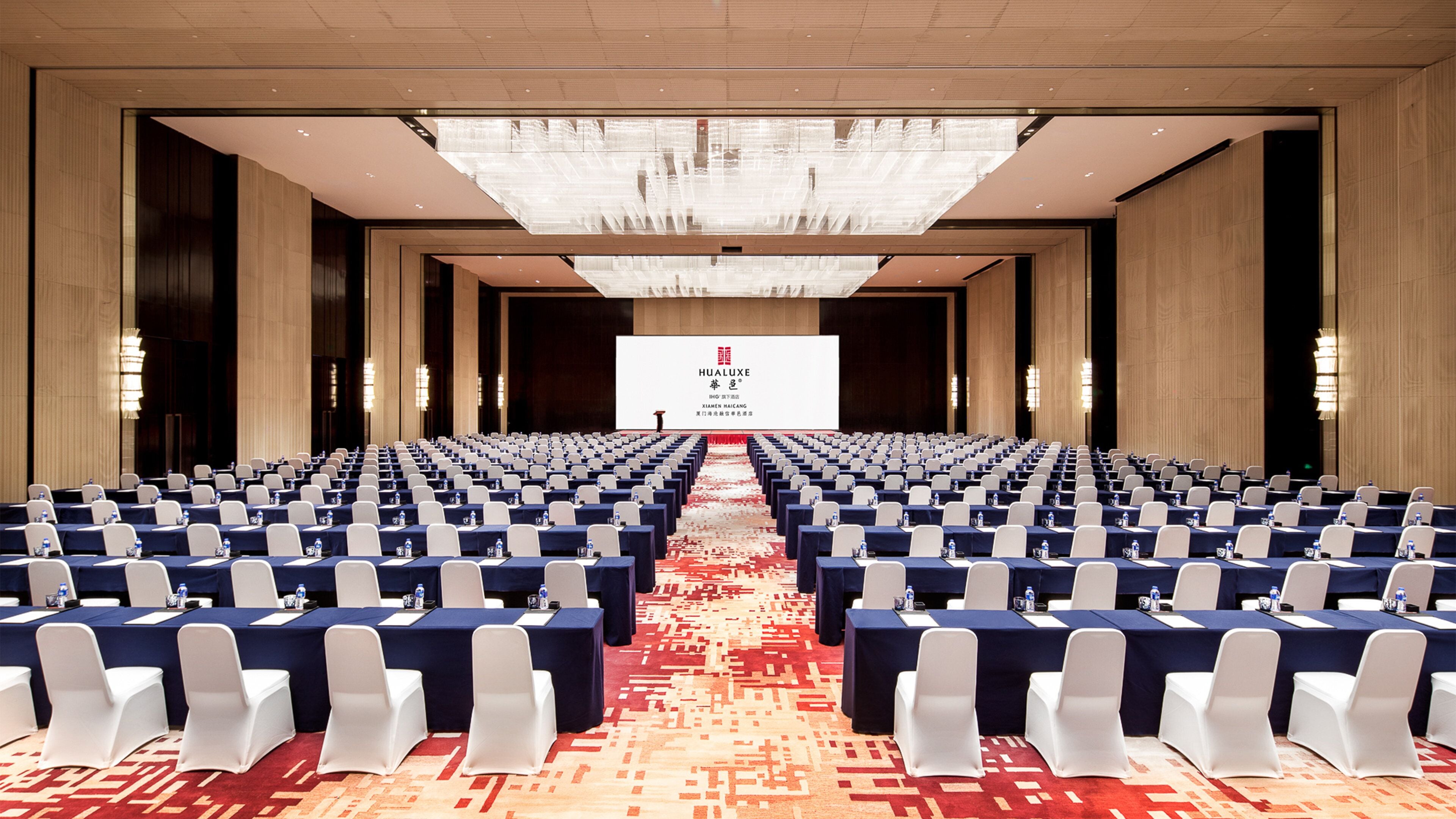 Un salón de conferencias con filas de mesas y sillas, suelo alfombrado y una pantalla grande al frente.
