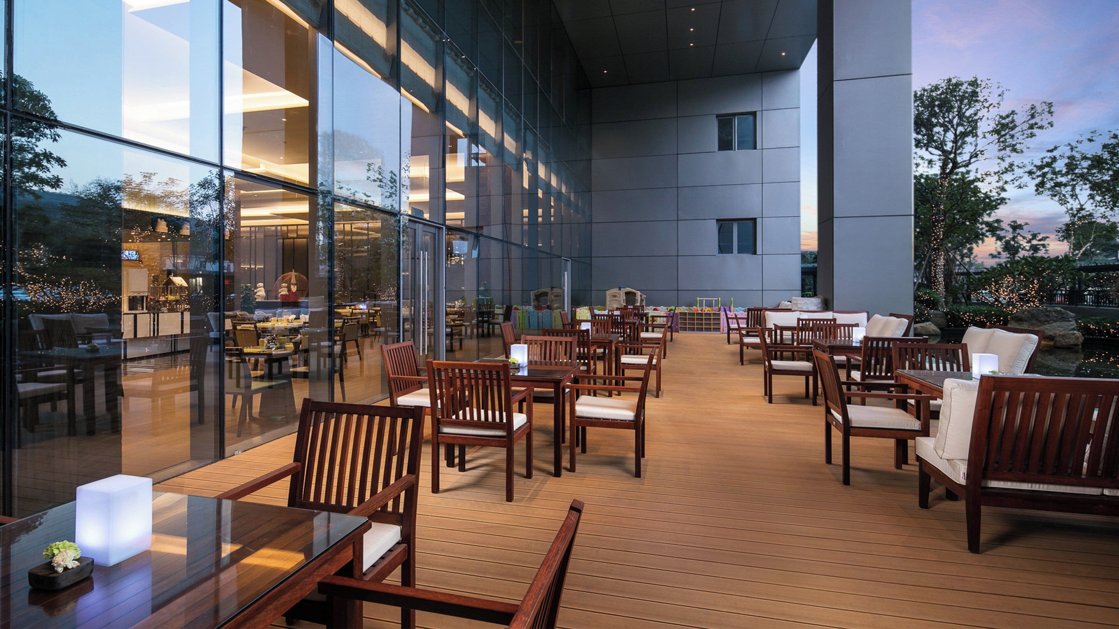 An outdoor dining area with wooden tables and chairs, large windows, and a view of the interior dining space.