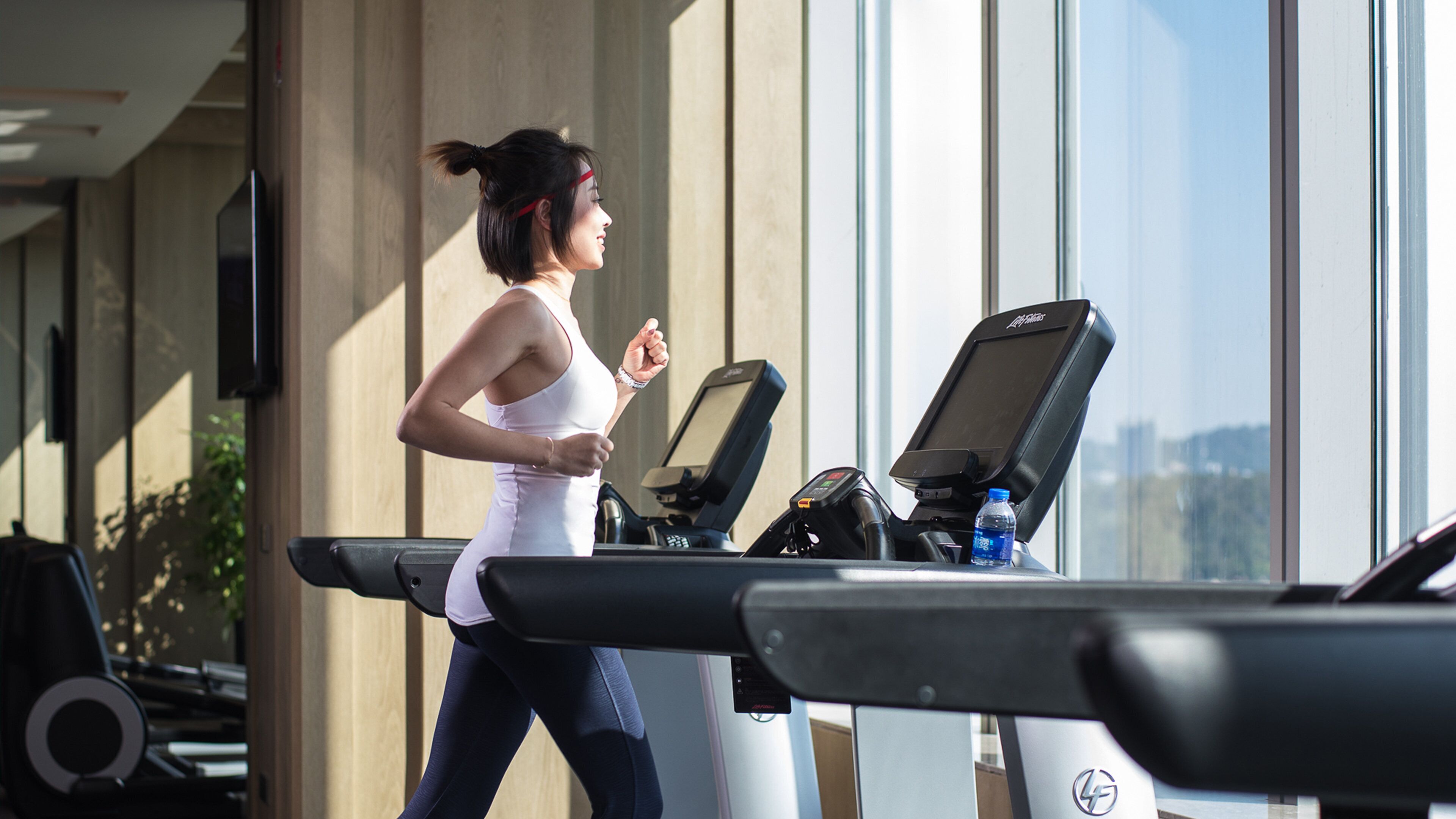 Una mujer corre en una cinta de correr en un gimnasio con ventanas amplias y equipamiento de ejercicio.