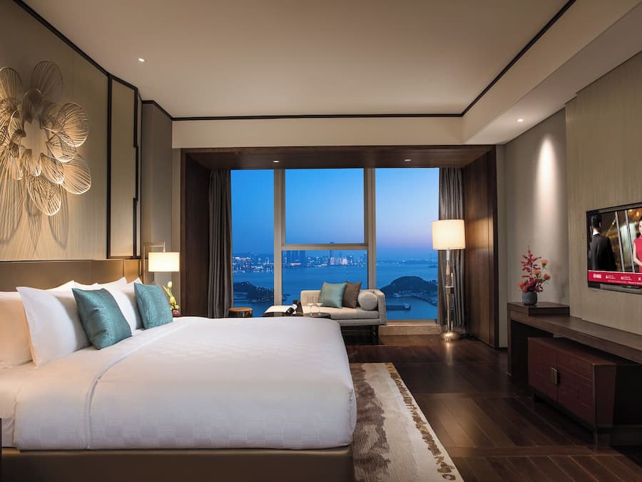 A modern hotel room with a large bed, a television, and a view of a cityscape through large windows.