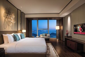 A modern hotel room with a large bed, a television, and a view of a cityscape through large windows.