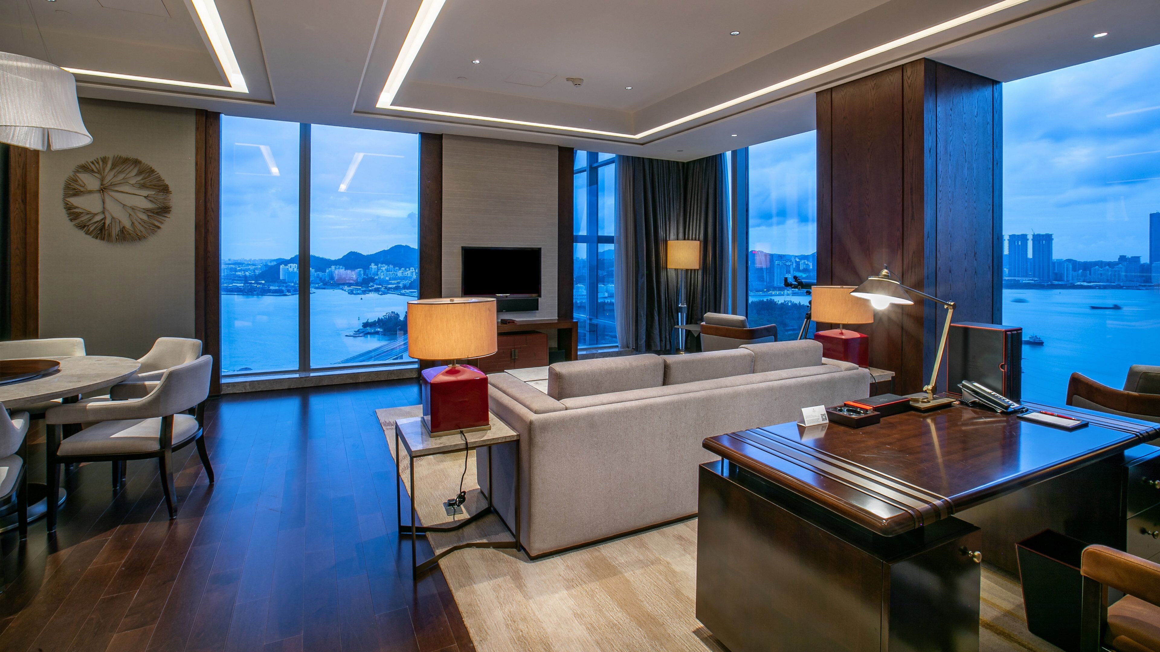 A modern hotel room with a large window offering a city view, a sofa, a dining area, and a desk with a lamp.