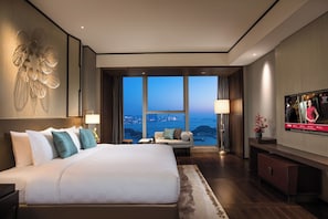 A modern hotel room with a large bed, a television, and a view of a cityscape through large windows. - HUALUXE Xiamen Haicang Harbour View by IHG (Xiamen)