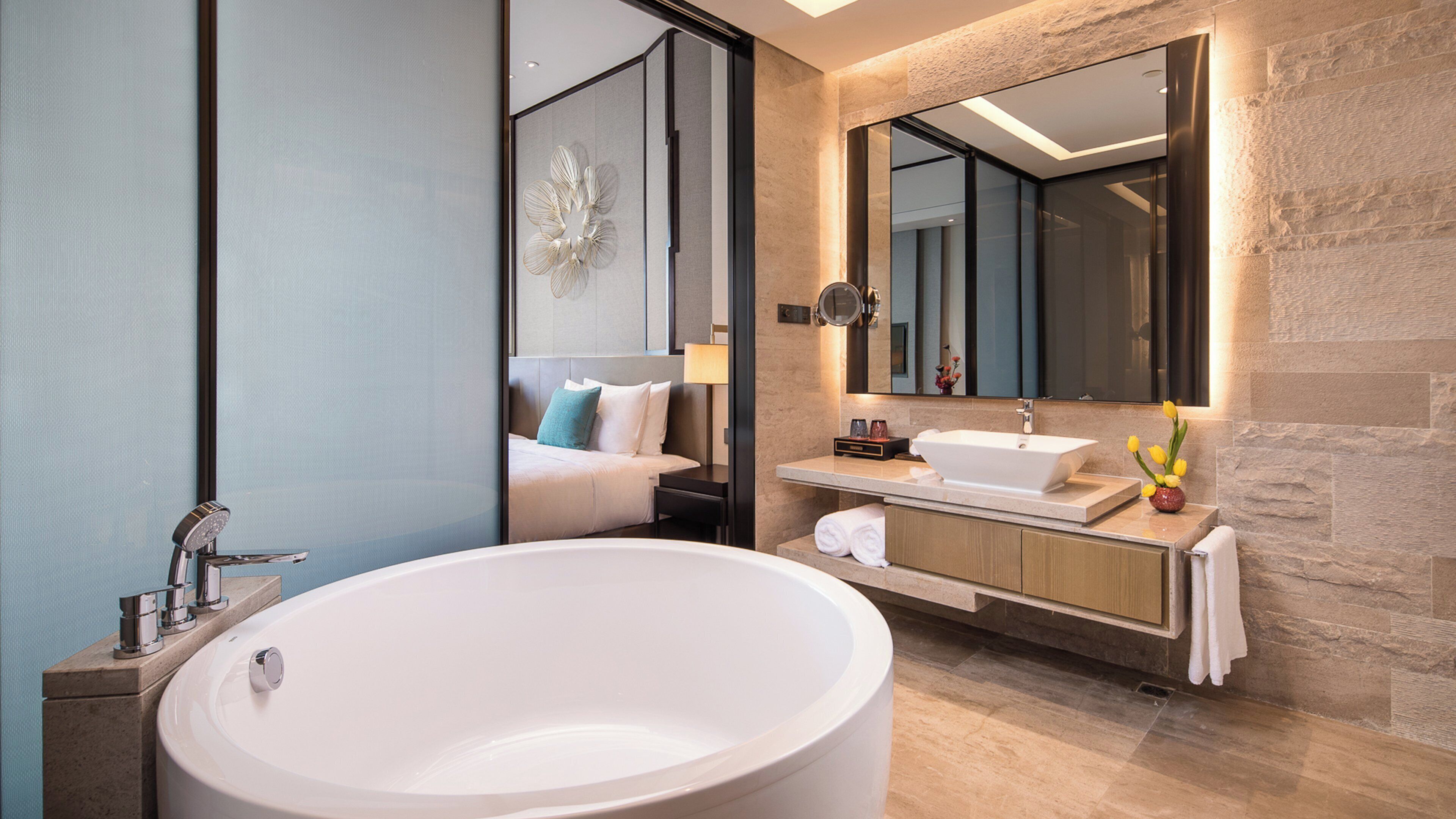 Une salle de bain moderne avec une baignoire sur pieds, un meuble-lavabo avec miroir et un lit visible à travers une cloison en verre.