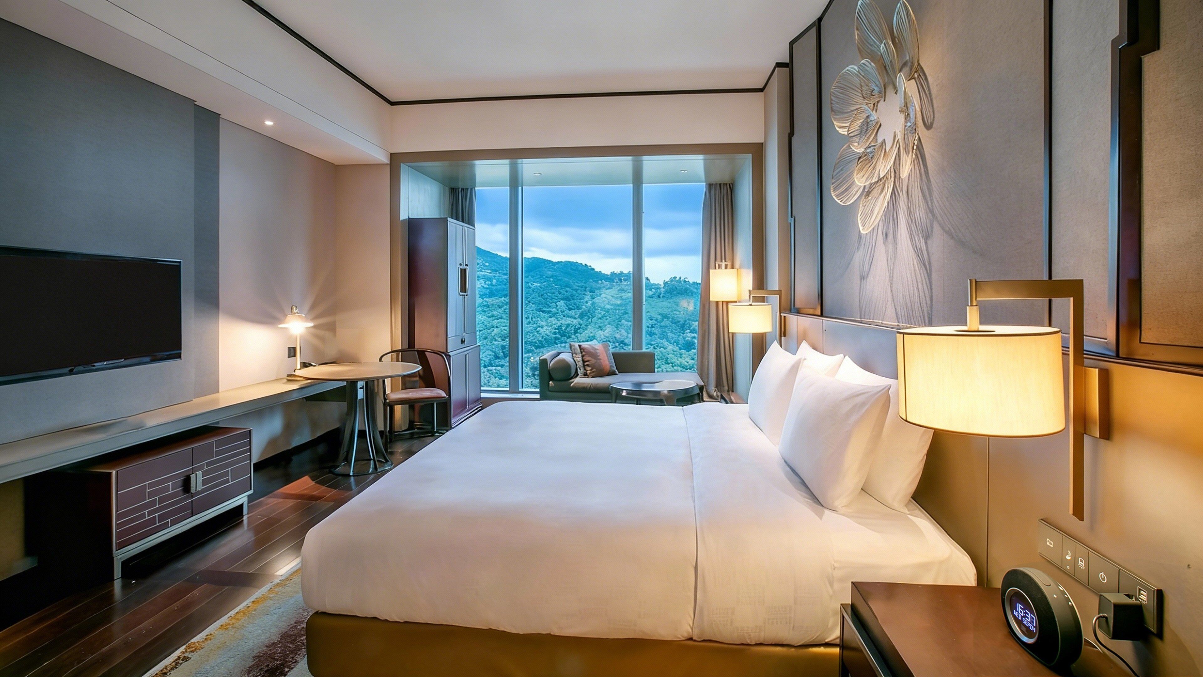 A modern hotel room with a large bed, a TV, a desk, and a view of a mountainous landscape.